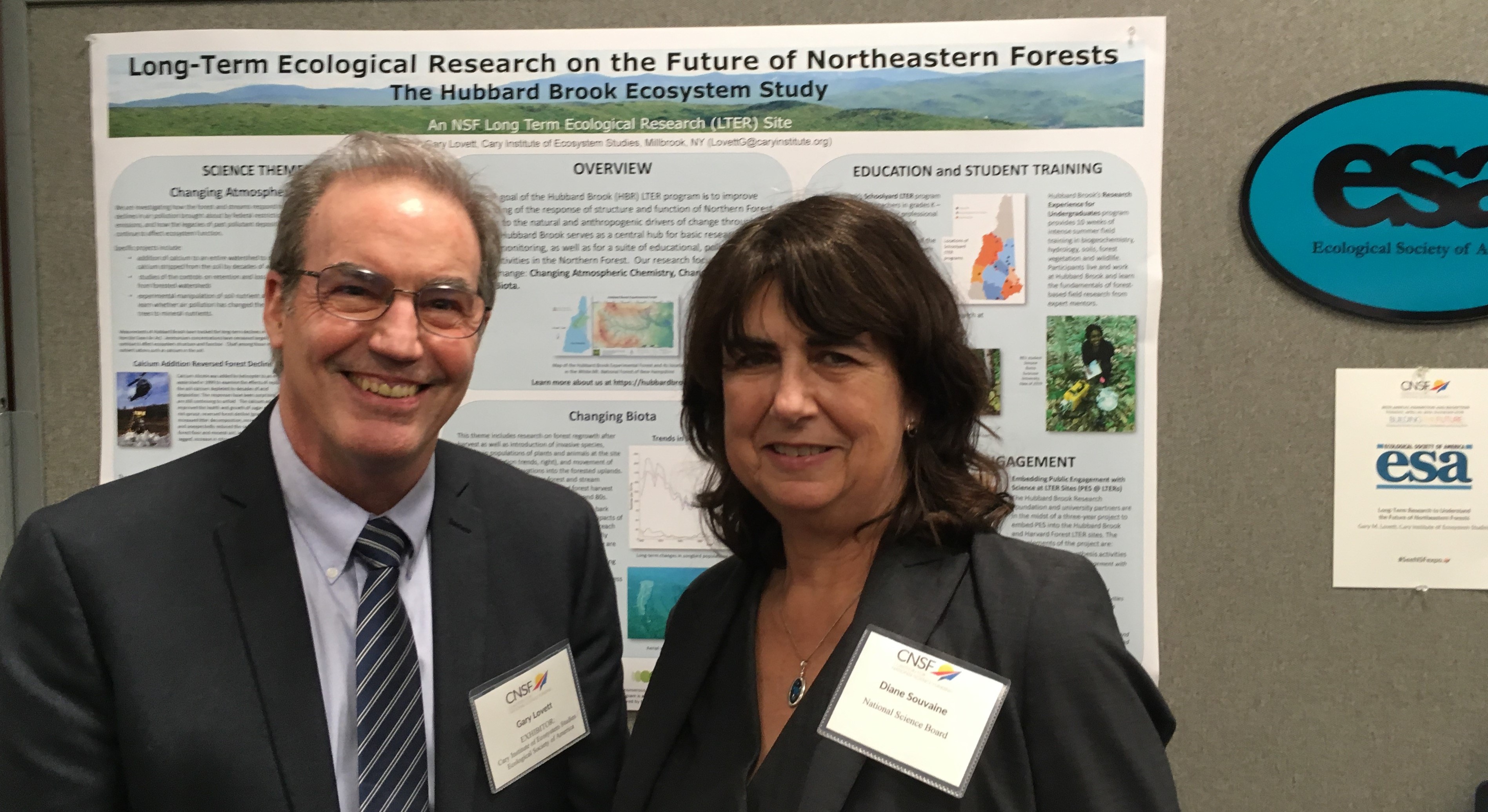 Two people smile before the camera in front of a poster which is titled 'long-term ecological research on the future of northeastern forests'