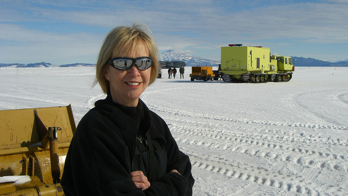 A scientist stands on an icy plain in front of large yellow equipment